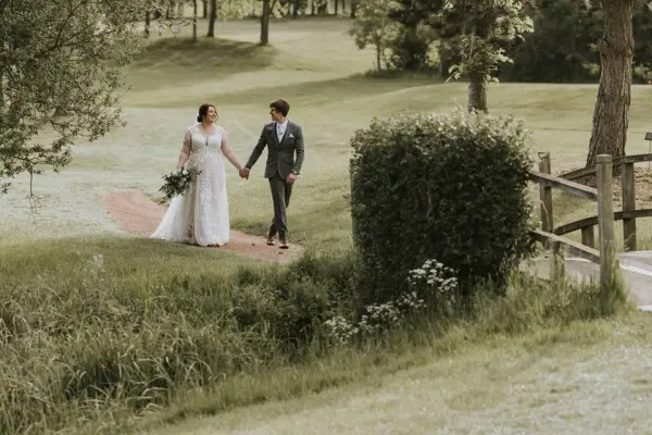 A couple walks hand-in-hand along a scenic path, surrounded by lush greenery and rustic fencing, perfect for an outdoor wedding celebration.