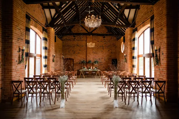 Rustic ceremony space with exposed brick walls, wooden beams, and elegant chandelier, featuring rows of crossback chairs and floral accents.