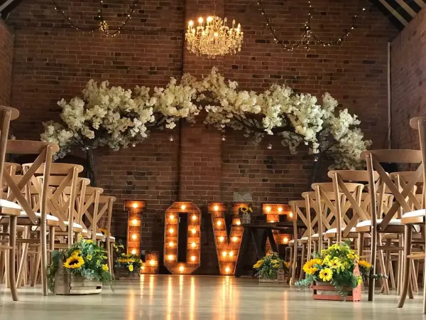Intimate ceremony space featuring charming wooden chairs, a floral arch, and lit "LOVE" signage against a rustic brick backdrop.