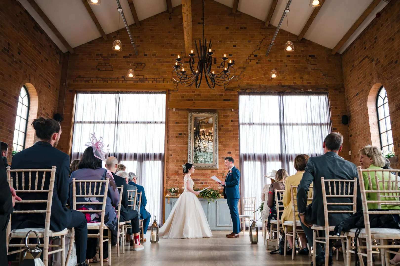 Ceremony in a rustic venue featuring exposed brick walls and large windows, with guests seated on chiavari chairs, creating an intimate atmosphere.