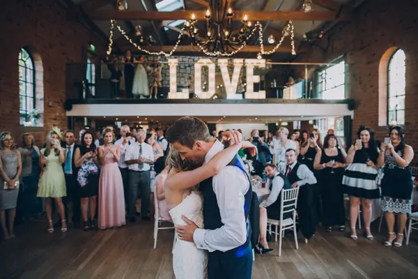 Couple shares a romantic first dance in a rustic venue with exposed brick walls, wooden beams, and elegant "LOVE" marquee lights.
