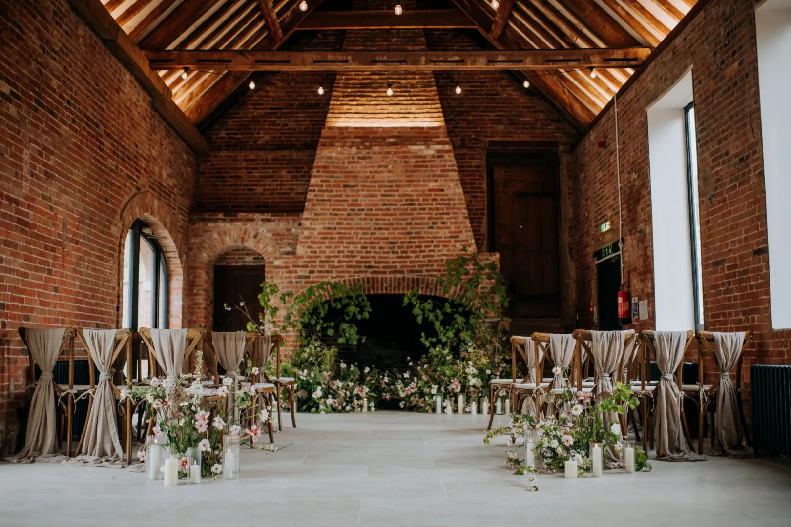 Rustic wedding venue featuring exposed brick walls, elegant chairs adorned with flowing fabric, and lush floral arrangements creating a romantic ceremony space.