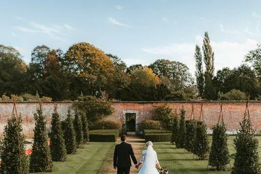 Couple walks hand-in-hand through a manicured garden, framed by lush hedges and a charming brick wall in the background.
