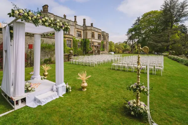 Beautiful outdoor ceremony space featuring a decorated arbor, elegant gold accents, and white chairs set on lush green grass.