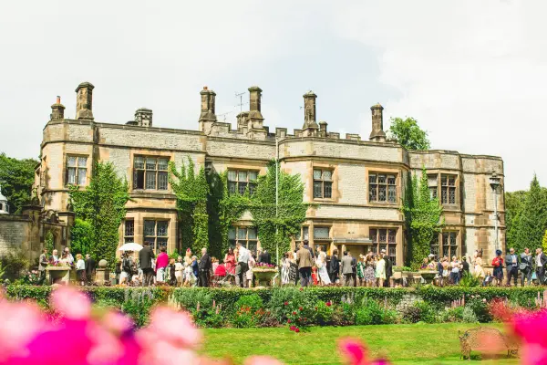 Elegant stone mansion surrounded by lush gardens, with guests mingling in the sun and vibrant flowers framing the scene.