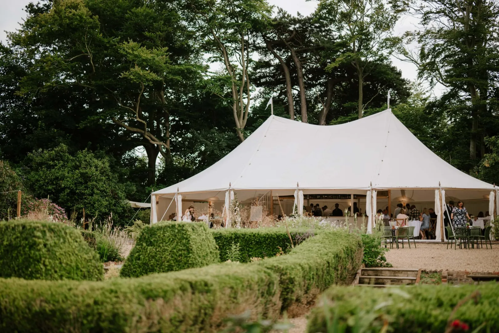 Charming outdoor wedding venue featuring a large white tent surrounded by lush greenery and beautifully manicured hedges.
