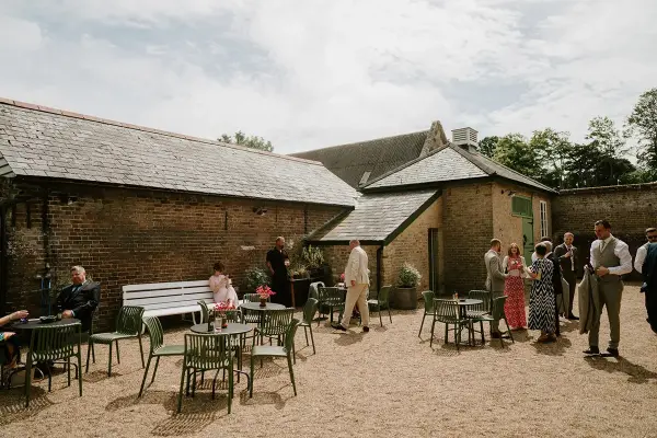 Charming outdoor courtyard with gravel flooring, rustic brick walls, and green seating, ideal for mingling during a wedding reception.