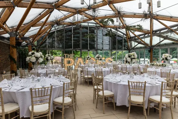 Bright, airy reception space featuring elegant round tables set for dining, surrounded by lush floral arrangements and a stunning glass roof.