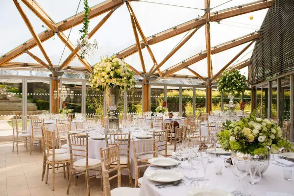 Stylish indoor reception with wooden beams, elegant tables adorned with floral arrangements, and ample natural light streaming through glass walls.