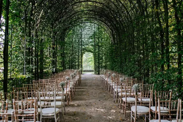 Charming outdoor wedding ceremony space with an elegant green archway, lined with rustic chairs and surrounded by lush foliage.