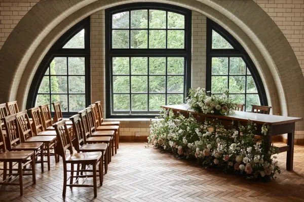 Bright indoor ceremony space featuring natural light through large windows, wooden chairs, and a floral-adorned altar table.