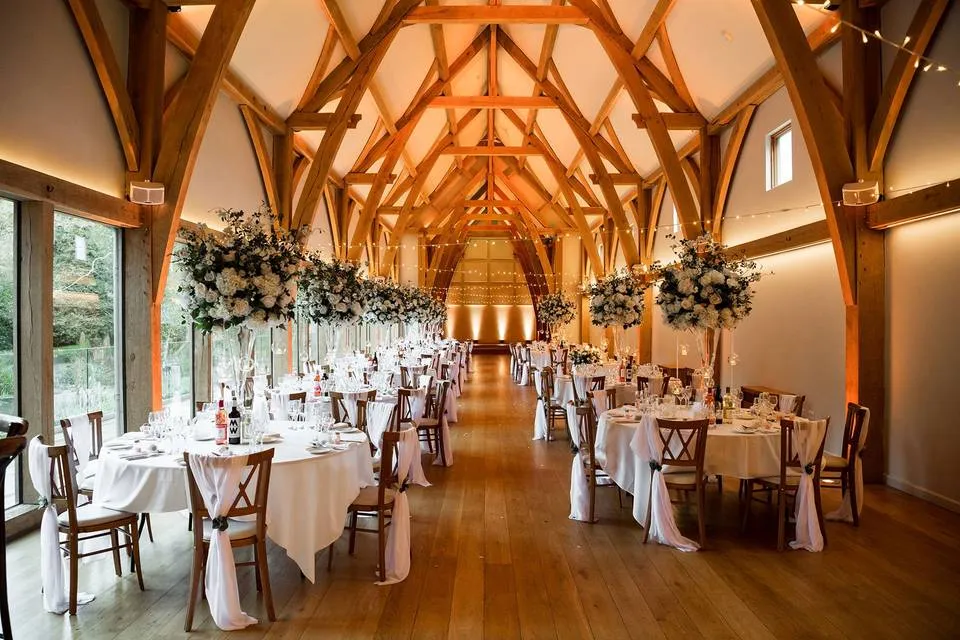 Elegant reception hall featuring tall wooden beams, spacious tables adorned with floral centerpieces, and warm ambient lighting.