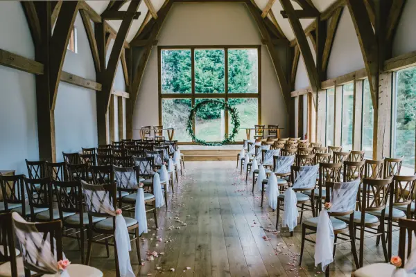 Charming indoor ceremony space with wooden beams, elegantly arranged chairs, and a beautiful greenery backdrop through large windows.