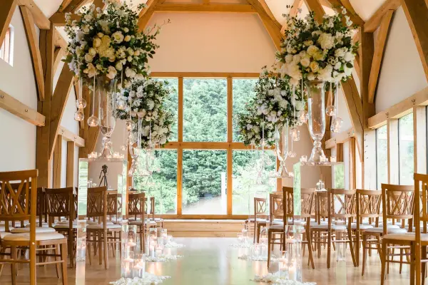 Bright rustic indoor ceremony space featuring elegant wooden beams, hanging floral arrangements, and a stunning large window overlooking greenery.