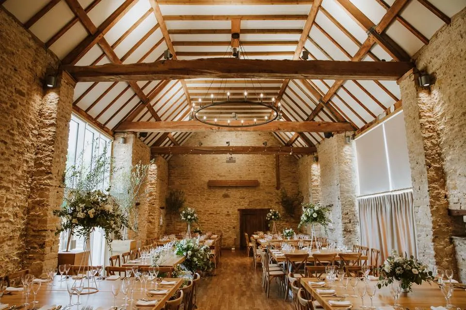 Rustic barn venue featuring high wooden beams, elegant chandeliers, and long tables adorned with floral centerpieces. Natural light floods the space.