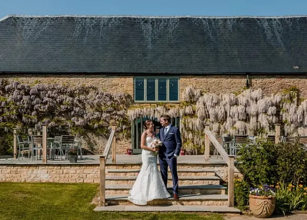 A bride and groom pose on a wooden staircase in front of a rustic barn, adorned with blooming wisteria and vibrant greenery.
