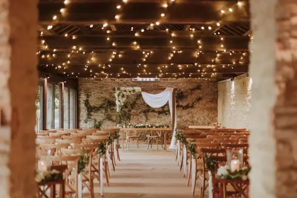 Charming indoor ceremony space featuring rustic stone walls, warm string lights, and wooden chairs adorned with greenery.