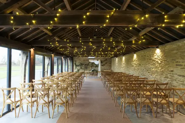 Charming rustic barn featuring wooden cross-back chairs, twinkling fairy lights, and natural stone walls for a cozy ceremony setting.