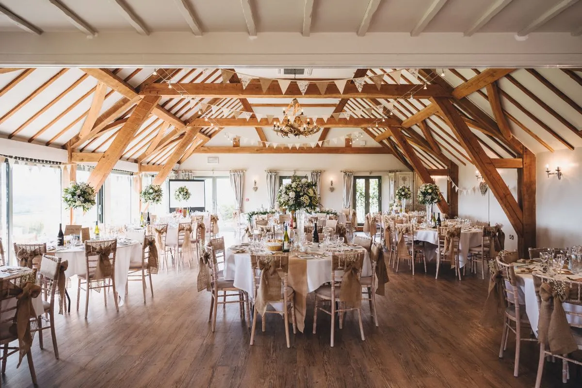 Spacious reception area featuring rustic wooden beams, elegantly set tables, and beautiful floral arrangements for a charming wedding atmosphere.