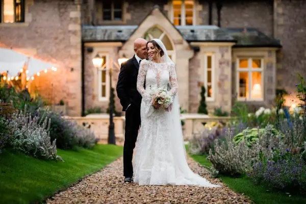 A bride and groom share an intimate moment on a gravel path surrounded by lush gardens and a charming stone mansion. Soft lighting enhances the romantic vibe.