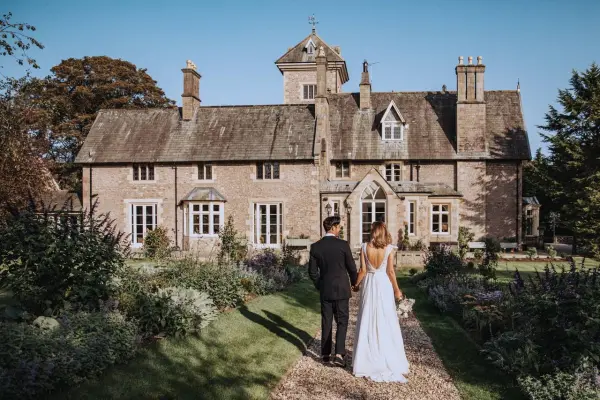 A couple walks hand-in-hand toward a charming stone mansion surrounded by lush gardens, perfect for a romantic outdoor wedding.