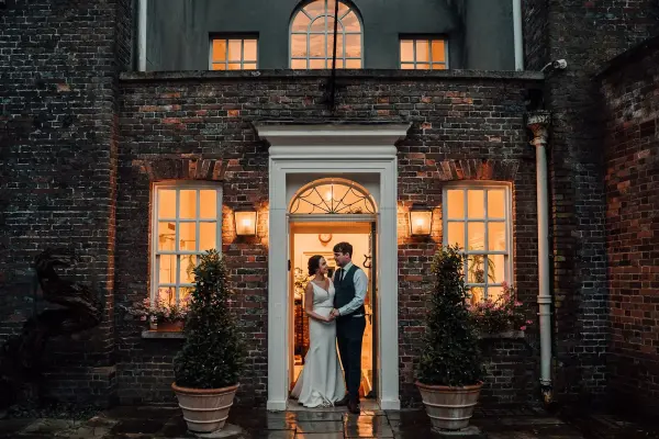 Couple standing at an elegant entrance with glowing windows, flanked by potted greenery, exuding a romantic ambiance ideal for weddings.
