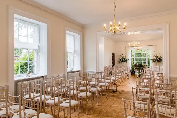 Bright, elegant ceremony space with white walls, large windows, and rows of chiavari chairs set for a wedding, complemented by floral decor.