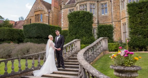 Couple poses on a curved stone staircase, with a majestic historical venue and manicured gardens in the background, featuring vibrant flowers.