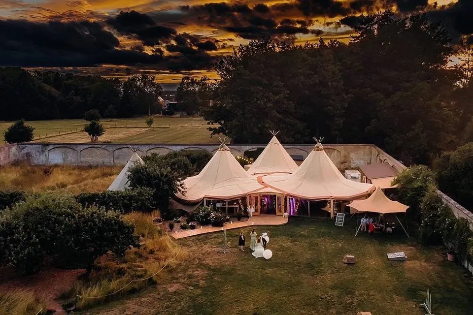 Three elegant tipis nestled in a lush green garden, surrounded by trees, under a dramatic sunset sky, perfect for outdoor weddings.