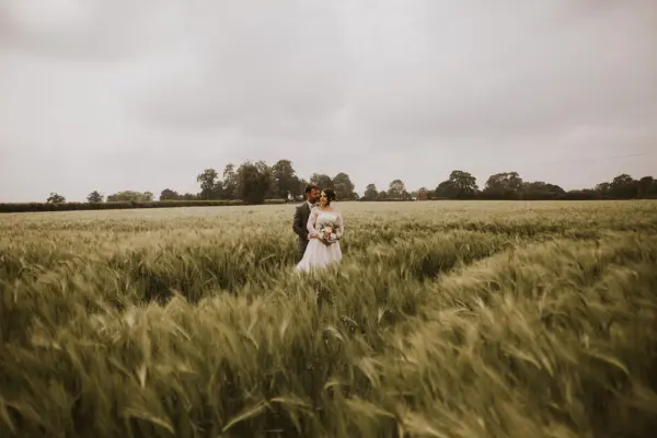 A couple stands in a lush green field, surrounded by tall grass under a soft, cloudy sky, creating a romantic outdoor wedding setting.