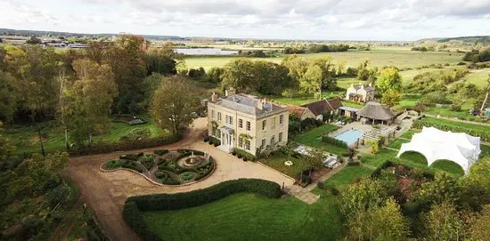 Aerial view of a charming mansion surrounded by manicured gardens, a serene pool, and a spacious outdoor event tent. Lush greenery enhances the scene.