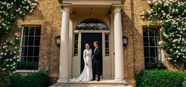 A couple stands elegantly on the steps of a grand entrance, flanked by lush greenery and blooming flowers, perfect for a romantic ceremony.