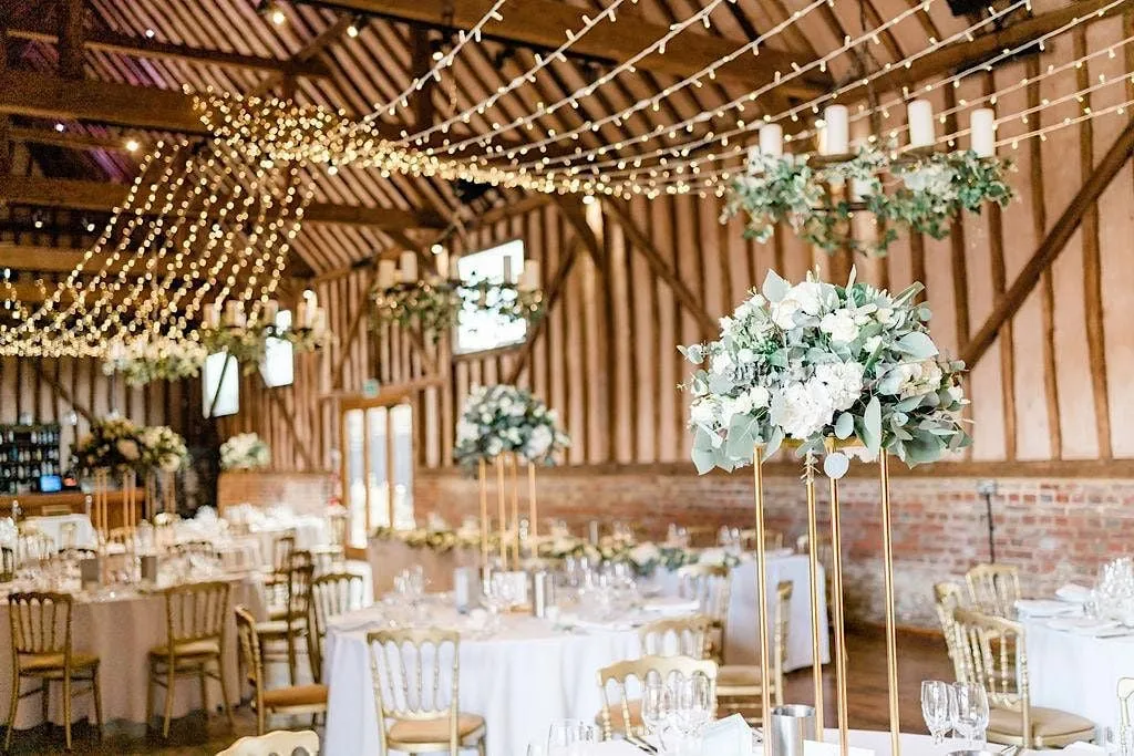 Rustic barn interior featuring elegant floral centerpieces, gold chairs, and string lights across wooden beams, creating a romantic atmosphere.