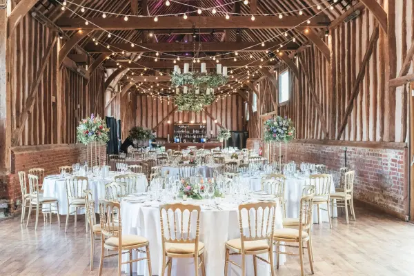 Rustic barn venue featuring elegant round tables with floral centerpieces beneath warm string lights and wooden beam architecture.
