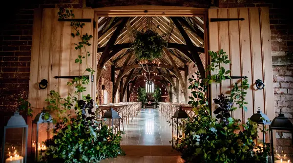Rustic barn entrance featuring wooden doors, lush greenery, and a charming ceremony space with elegant chandeliers and rows of chairs.