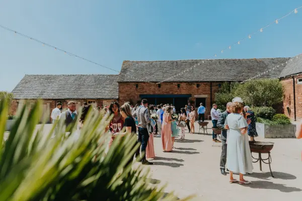 Guests gather in a spacious outdoor area with rustic brick buildings, string lights, and lush greenery, perfect for a charming wedding celebration.