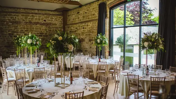 Elegant reception space featuring round tables adorned with tall floral centerpieces and large windows, showcasing natural light and rustic stone walls.