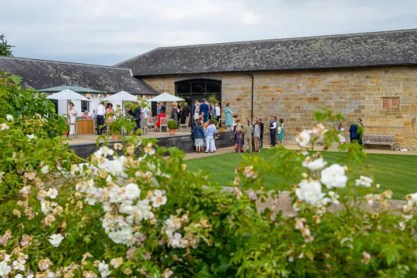 Guests mingle outside a rustic stone barn, adorned with blooming flowers and shaded seating areas, ideal for a charming outdoor celebration.