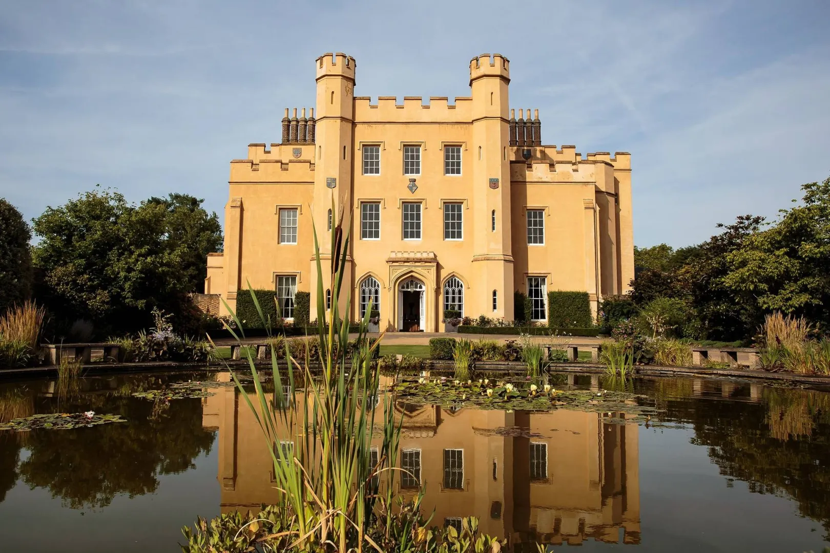 Sunlit castle with turrets surrounded by lush greenery and a tranquil pond reflecting its elegant facade, perfect for a romantic wedding.