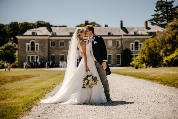 A bride and groom kiss on a gravel path, framed by a historic mansion and lush greenery, ideal for elegant outdoor weddings.
