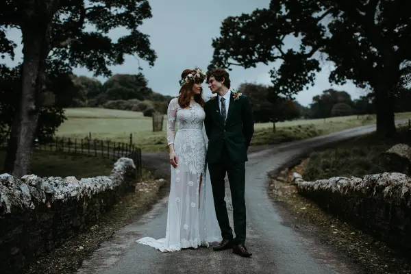 A couple stands together on a picturesque country road, surrounded by lush greenery and rustic stone fences, perfect for a romantic wedding setting.