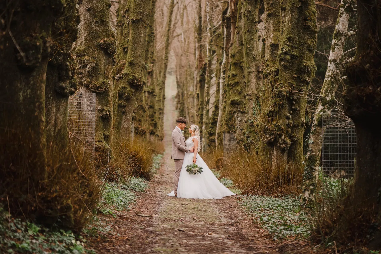 A romantic couple embraces along a tree-lined path, surrounded by lush greenery and rustic forest charm, perfect for a woodland wedding.