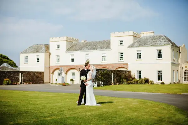 A couple embraces on lush green lawn in front of a charming estate with castle-like architecture, perfect for an elegant wedding.