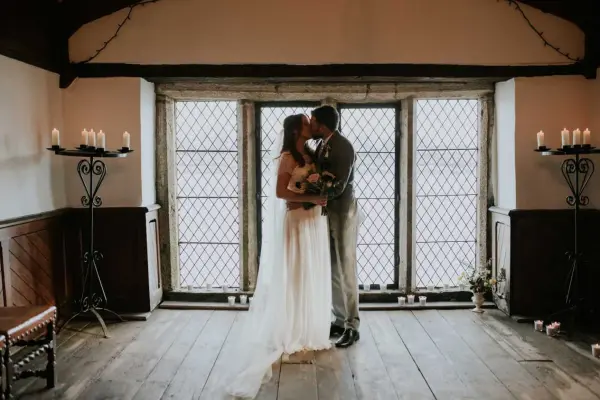 Couple sharing a kiss in a charming indoor space, featuring large windows, wooden floors, and elegant candle stands.