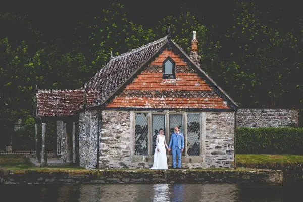 Charming stone cottage by a serene lake, featuring a couple in elegant attire standing together, surrounded by lush greenery.