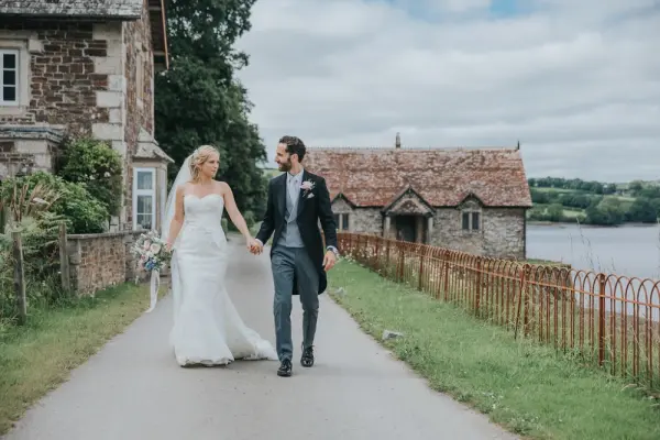 Elegant couple walking hand-in-hand along a scenic path by a rustic venue near a lake, framed by lush greenery and charming architecture.