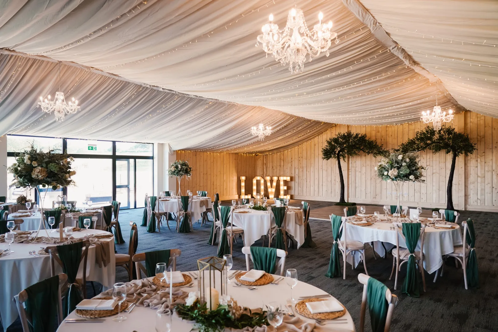 Elegant reception hall featuring draped ceilings, crystal chandeliers, and beautifully arranged tables with greenery and a "LOVE" display.