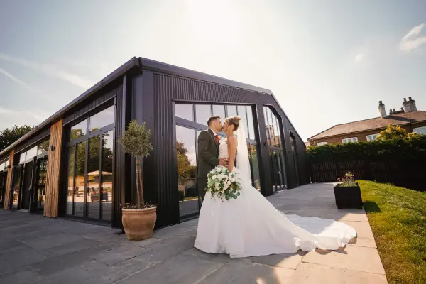 Couple embraces outside a modern venue with large windows, surrounded by greenery, ideal for a romantic outdoor ceremony or elegant reception.