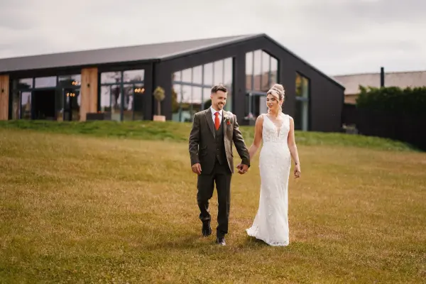 Elegant couple walking hand in hand on a grassy lawn, with a modern venue featuring large windows in the background, perfect for outdoor ceremonies.