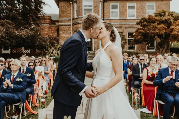 A bride and groom share a kiss during their outdoor ceremony, surrounded by guests seated in elegant attire amidst lush greenery.